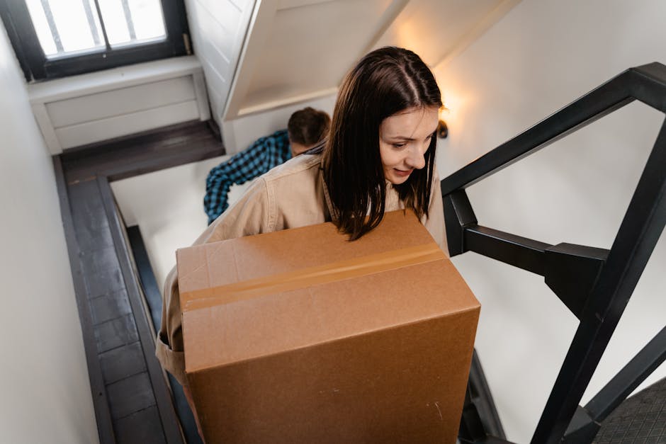 A woman with shoulder-length brown hair is carrying a large cardboard moving box up a narrow staircase inside a residential property, guided by natural ambient lighting. She is wearing a beige jacket