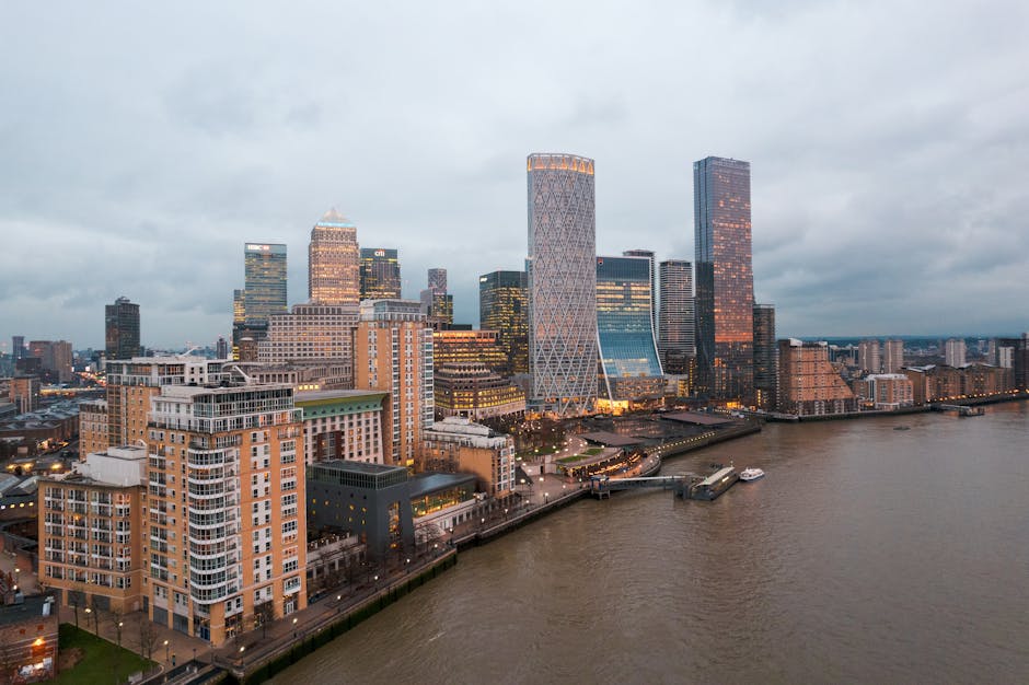 A panoramic view of London's Canary Wharf skyline during early evening, showcasing tall modern office buildings constructed with glass and steel, illuminated with internal and external lighting. In th