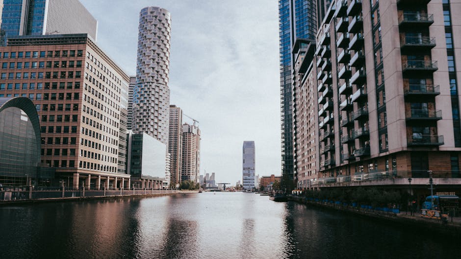 A panoramic view of London's Canary Wharf skyline during early evening, showcasing tall modern office buildings constructed with glass and steel, illuminated with internal and external lighting. In th